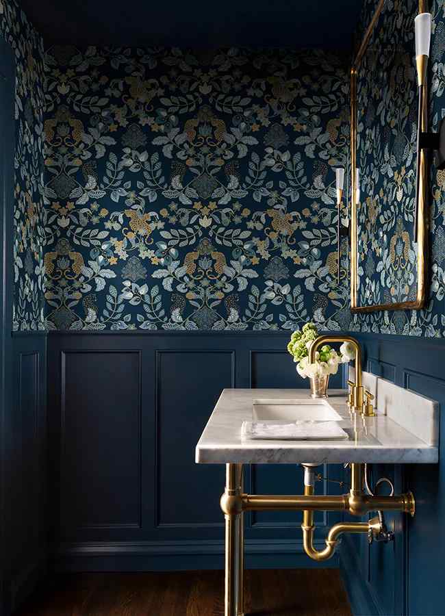 A navy bathroom with wallpaper and wooden floor and gold fixtures