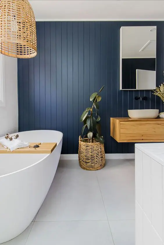A navy blue wooden panel wall and white bathtub and woven plant basket